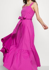 Fuksia värinen hihaton mekko, jossa on neliönmuotoinen pääntie, vyötärönauha, leveä helma ja sileä kangas sekä röyhelöinen alaosa.