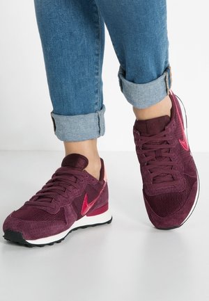 Pieds portant un jean bleu retroussé et des baskets Nike bordeaux avec des logos swoosh rouges sur un fond blanc.