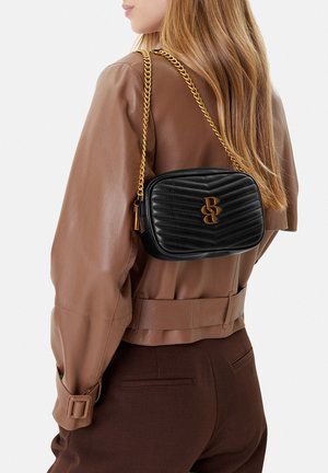 Frau trägt braune Lederjacke und Hose, trägt eine schwarze gesteppte Umhängetasche mit goldener Kettenriemen und goldenem Logo.