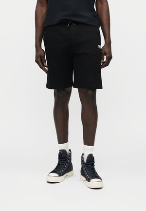 Férfi fekete zsinóros rövidnadrágban, fehér zokniban és fekete magas szárú tornacipőben, egyszínű háttér előtt.