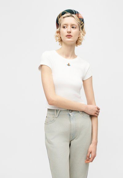 Ξανθιά γυναίκα που φοράει λευκό εφαρμοστό κοντομάνικο τοπ με μικρό πολύχρωμο έμβλημα, ανοιχτό γκρι τζιν και καρό μαντήλι στο κεφάλι, στέκεται με τα χέρια σταυρωμένα.