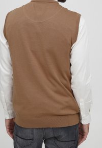 Brunstickad väst med ribbad fåll, bärs över en vit långärmad skjorta. Baksidan har en slät textur och en rundad halsringning.