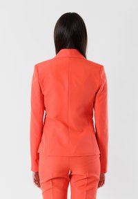 Blazer corallo; vestibilità sartoriale con colletto a scollo a V, texture liscia, due cuciture posteriori e pantaloni abbinati. Design minimalista, senza decorazioni visibili.