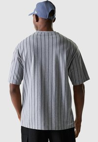 Homme portant une chemise à manches courtes gris à rayures et une casquette de baseball bleue, tourné le dos, se tenant devant un fond clair uni.