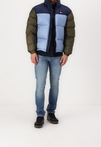 Péřová bunda v námořnické modři, světle modré a zelené barvě. Přiléhavý střih s vysokým límcem, předním zipem a logem. Nošena s světlými džínami a černými teniskami.