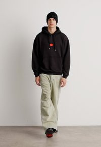 Felpa nera con un patch logo rosso, abbinata a jeans larghi verde chiaro. Il modello indossa un berretto nero con "HUGO" ricamato.
