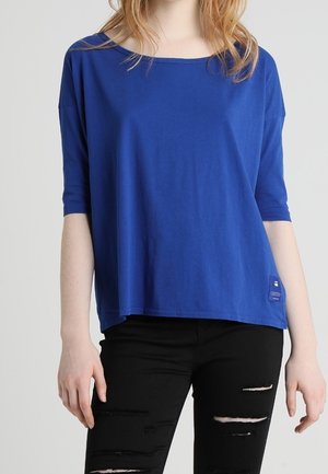 Frau trägt ein locker sitzendes royalblaues Kurzarmtop und schwarze, zerrissene Skinny-Jeans vor einfarbigem Hintergrund.