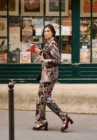 Ein gemustertes bordeauxrotes Anzug mit Details in Weiß und Blau, kombiniert mit passenden Schuhen mit Blockabsatz. Die Person trägt eine schwarze Clutch und eine Baguette-Tasche.