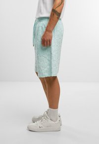 Lys teal teksturerede svøm shorts med snor i taljen, båret med hvide sneakers og korte sokker, set fra siden.