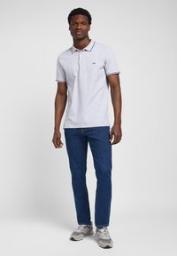 Camisa polo gris con cuello y acentos de mangas en contraste color navy, combinada con jeans de mezclilla azules y zapatillas grises. Conjunto casual simple y pulcro.