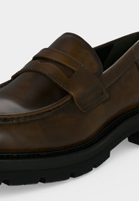 Καφέ δερμάτινο loafer με ραφή moc toe, λουράκι στο πάνω μέρος του ποδιού και χοντρή μαύρη καουτσούκ σόλα με πέλμα.