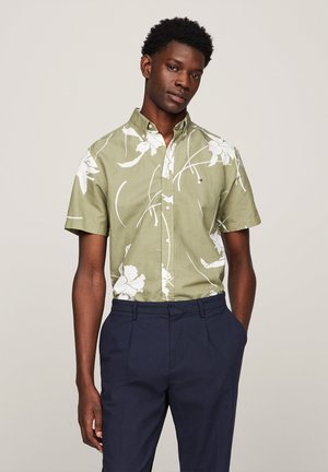 Camicetta a maniche corte con bottoni di colore verde oliva, con motivo floreale bianco. Abbinata a pantaloni blu scuro, su uno sfondo neutro.