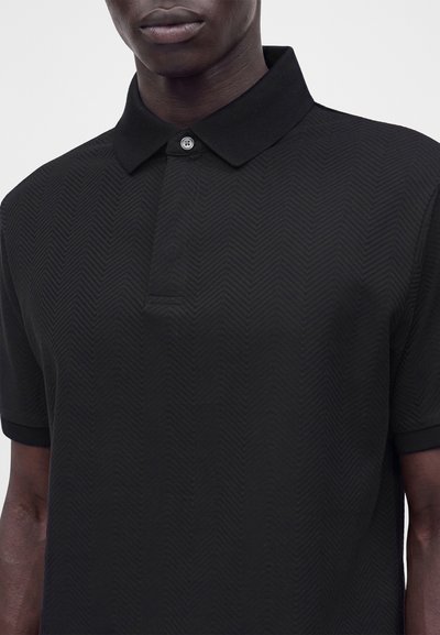 Gros plan sur une personne portant un polo noir texturé avec un col et un seul bouton fermé.