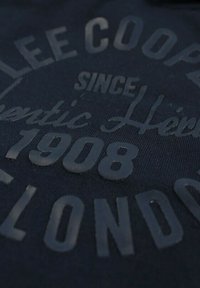 Tissu foncé avec texte en relief indiquant « Lee Cooper Depuis 1908 Londres » dans un design circulaire.