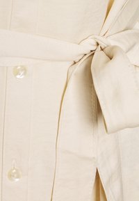 Camicia beige con cintura a cravatta, chiusura con bottoni e superficie liscia con cuciture visibili lungo i bordi.