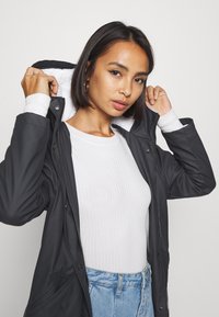 Svart vanntett jakke med hette foret med faux pels, glidelås foran og sidelommer. Modellen har på seg en hvit ribbestrikket genser og høytlivsjeans.