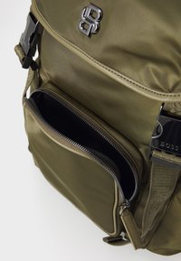 Olive zeleni nylon nahrbtnik z glavnimi predelom in sprednjim žepom na zadrgo. Ima srebrne dodatke s podrobnostmi logotipa in teksturirane naramnice.