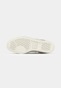 Sneaker outsole featuring a textured beige rubber material, a herringbone pattern, and a central logo for traction and grip.