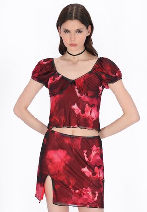 Młoda kobieta w czerwonym kwiecistym topie z bufkami i dopasowanej spódnicy z bocznym rozporkiem, pozująca z rękami na biodrach na białym tle.