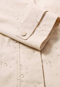 Veste imperméable beige avec des gouttelettes d'eau sur le tissu. Dispose d'un poignet à boutons et de coutures visibles. Texture lisse et design contemporain.