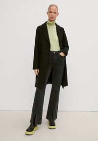 Black long coat over a light green turtleneck, paired with high-waisted dark jeans featuring side slits and chunky black and neon green boots.