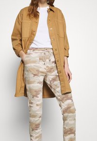 Chemise boutonnée marron clair, t-shirt blanc en dessous, et pantalon beige camouflage avec taille à cordon. Tissu doux, coupe décontractée, et poches visibles.