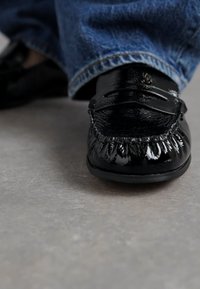 Zwarte leren moccasin schoen met een gestructureerde afwerking en gedetailleerde stiksels, gedragen met blauwe spijkerbroeken op een grijze ondergrond.