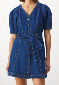 Denimmekko V-pääntiellä, lyhyillä puffihihnoilla ja istuvalla vyötäröllä. Varustettu nappikiinnityksellä edessä ja vastaavalla kankaisella vyöllä. Tumma sininen väri.