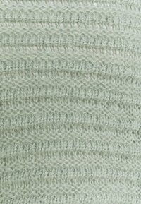 Zbliżenie jasnozielonej dzianiny o miękkiej, puchatej fakturze i naprzemiennych poziomych wzorach ściegów.