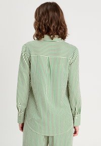 Chemise à manches longues rayée verte et blanche, avec des poignets boutonnés et un ourlet arrondi. Le tissu semble léger avec une texture lisse.
