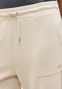 Κοντινό πλάνο μπεζ φόρμας με ελαστική μέση και κορδόνι, καθώς και μεγάλο μπροστινό τσέπη με μικρό χρυσό λογότυπο.