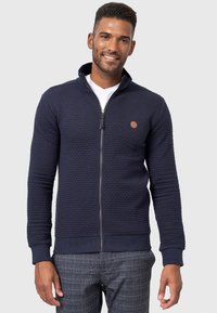 Mørkeblå zippered sweater med et struktureret mønster, ribbede manchetter og et lille logo-patch. Båret over en hvid T-shirt, sammen med ternede bukser.