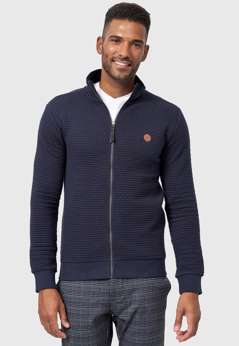 Mørkeblå zippered sweater med et struktureret mønster, ribbede manchetter og et lille logo-patch. Båret over en hvid T-shirt, sammen med ternede bukser.