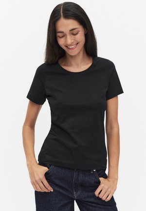 Vrouw glimlachend met gesloten ogen, gekleed in een eenvoudig zwart T-shirt met korte mouwen en donkerblauwe jeans, handen in de zakken.