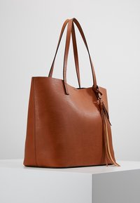 Bruine leren tote bag met een gestructureerde afwerking, twee lange handvatten en een decoratieve kwastdetail aan de zijkant. Minimalistisch ontwerp.