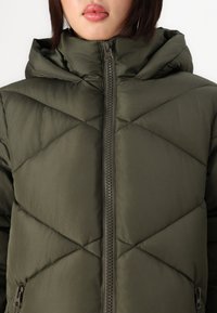 Doudoune vert olive avec fermeture éclair à l'avant, capuche et motif en losange matelassé. Elle dispose de poches latérales et d'une texture lisse et brillante.
