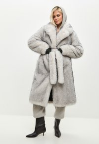 Palton gri din blană artificială, cu glugă, guler lat și curea asortată. Prezintă o textură cu nervuri și buzunare laterale. Poartă peste pantalonii gri și cizme negre.