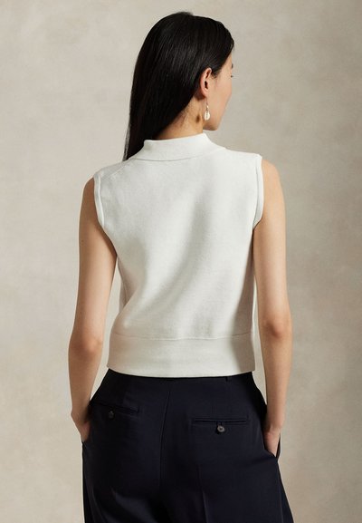 White sleeveless sweater with a collar, textured knit, and ribbed hem. Model wears dark pants with back pockets.