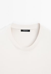 Jasnobeżowa koszulka z okrągłym dekoltem i żebrowanym wykończeniem przy szyi. Z przodu czarna metka z napisem "Massimo Dutti" oraz "Made in Portugal".