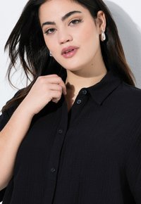 Czarna koszula z guzikiem, uszyta z teksturowanej tkaniny z kołnierzykiem, z ciemnymi guzikami. Ręka modela dotyka kołnierza, podkreślając detale wzornicze.