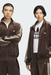 Brown velvet tracksuits with gold accents. The male model on the left wears a zip-up jacket, and the female model on the right sports a white T-shirt.
