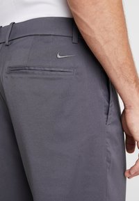 Närbild på en person som bär mörkgrå Nike-byxor, visar bakfickan med Nike-logotypen samt en del av höger arm och hand.