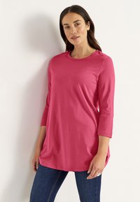 Vrouw met lang bruin haar, gekleed in een roze shirt met driekwart mouwen en een donkerblauwe spijkerbroek, staand tegen een witte muur met één hand ontspannen langs haar zijde.