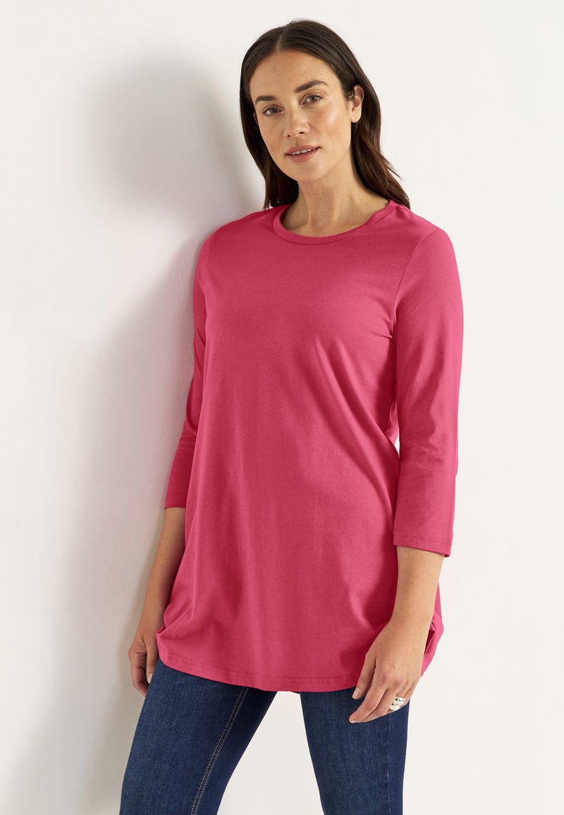 Vrouw met lang bruin haar, gekleed in een roze shirt met driekwart mouwen en een donkerblauwe spijkerbroek, staand tegen een witte muur met één hand ontspannen langs haar zijde.