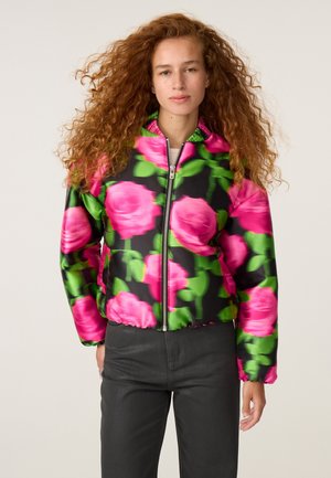 Vrouw met krullend rood haar die een zwarte jas draagt met grote roze rozen en groene bladpatronen en een zwarte broek, staand tegen een effen achtergrond.