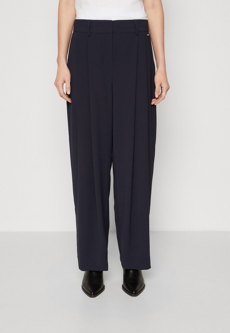 Femme vêtue d’un pantalon bleu marine taille haute et jambes larges, avec un top blanc et des bottines noires à bout pointu, debout sur un fond blanc.
