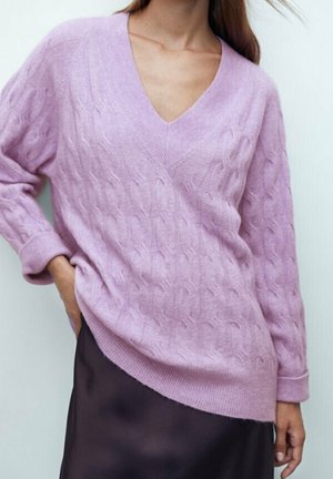 Femme portant un pull en maille torsadée violet clair à col en V et une jupe en satin foncé, avec une main dans la poche de la jupe.