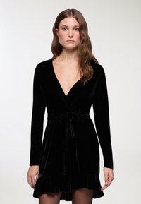Czarna aksamitna sukienka owinięta z długimi rękawami, głębokim dekoltem w serek, związaną w talii i falbaniastym dołem. Gładka tekstura, dopasowana sylwetka, minimalistyczny design.