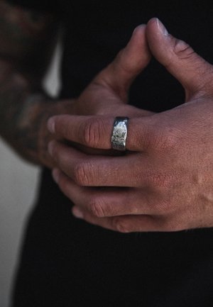 Silver ring with a textured surface, worn on a hand showing slight wrinkles and tanned skin. The background is dark with minimal detail.