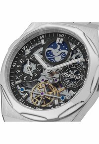 Montre automatique avec un boîtier en acier inoxydable argenté, cadran squeletté noir, fonction seconde zone horaire, mécanismes visibles et détail de logo.